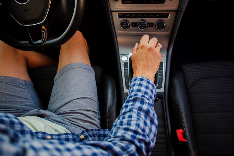 Vista de cima do interior de um carro, mostrando um motorista sentado no banco do condutor. Ele veste uma camisa xadrez azul e está com uma das mãos posicionada na alavanca de câmbio, sugerindo uma mudança de marcha ou ajuste do veículo. O volante aparece parcialmente à esquerda, enquanto o console central com controles e botões está em destaque. A cena transmite uma situação cotidiana de direção, com foco na interação do motorista com o veículo.
