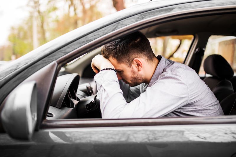 Homem sentado no banco do motorista apoia a cabeça no volante, demonstrando frustração ao tentar ligar o carro. A cena ocorre dentro do veículo, com o painel visível, e transmite uma situação comum de falha na partida. A imagem reforça o impacto de problemas mecânicos no dia a dia e a importância da manutenção preventiva para evitar esse tipo de imprevisto.