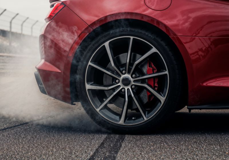Imagem em close de uma roda traseira de carro esportivo vermelho com fumaça saindo da região do freio, indicando aquecimento intenso durante a frenagem. O destaque para o disco e a pinça de freio reforça a importância do bom funcionamento do sistema para garantir segurança e evitar desgaste excessivo.