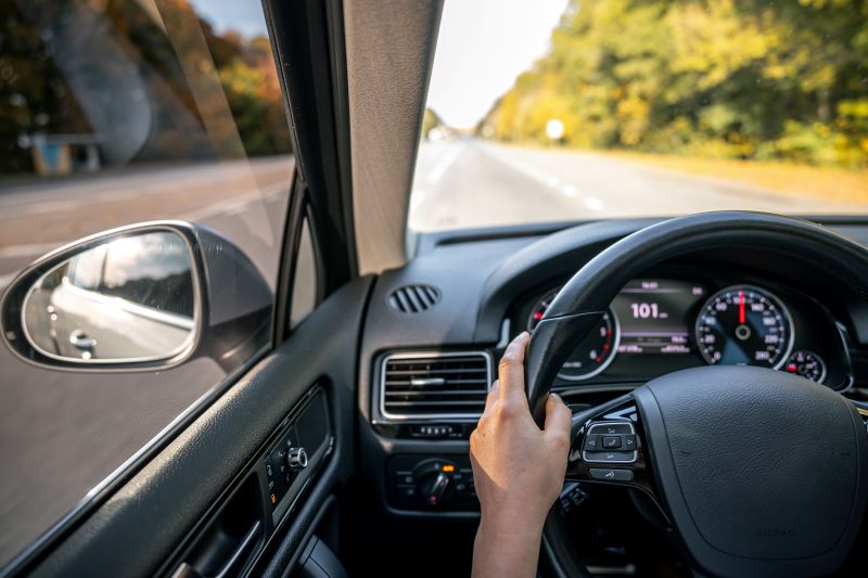 A imagem mostra a perspectiva de um motorista ao volante de um carro em movimento por uma estrada aberta. A mão do condutor segura o volante, enquanto o painel indica velocidade elevada. À frente, a estrada segue reta, cercada por árvores com tons esverdeados e amarelados, sugerindo um cenário de outono ou fim de verão. A luz natural é intensa, iluminando o interior do veículo e destacando os detalhes do painel e do retrovisor lateral, que reflete parte da estrada atrás. A cena transmite sensação de movimento, liberdade e viagem.